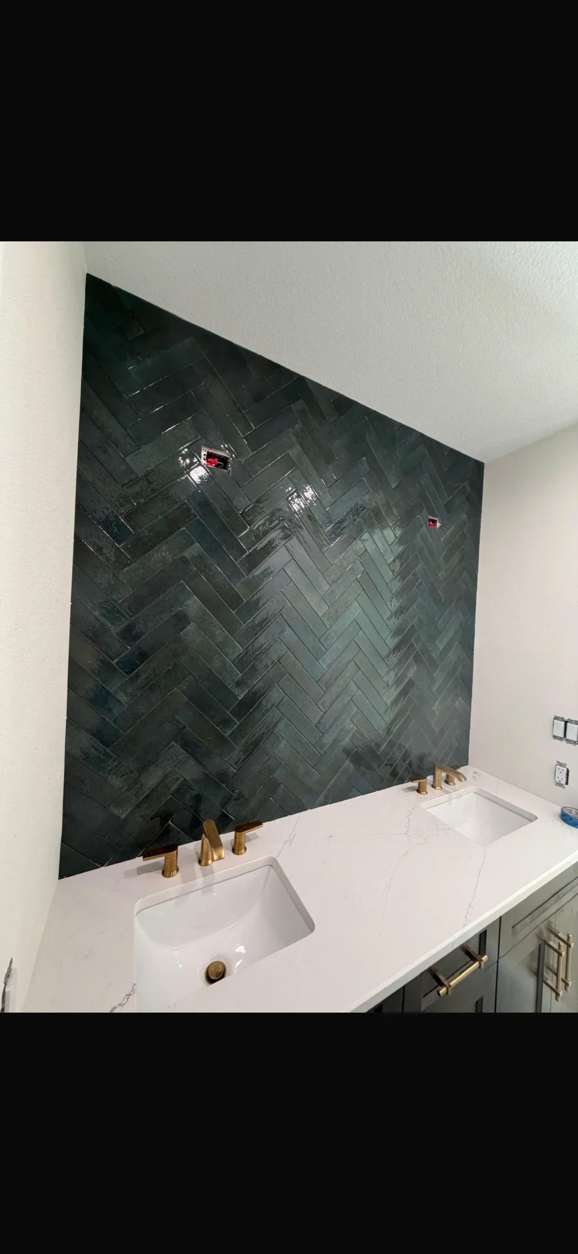 Charcoal gray herringbone tile backsplash behind bathroom vanity with dual sinks and brass fixtures