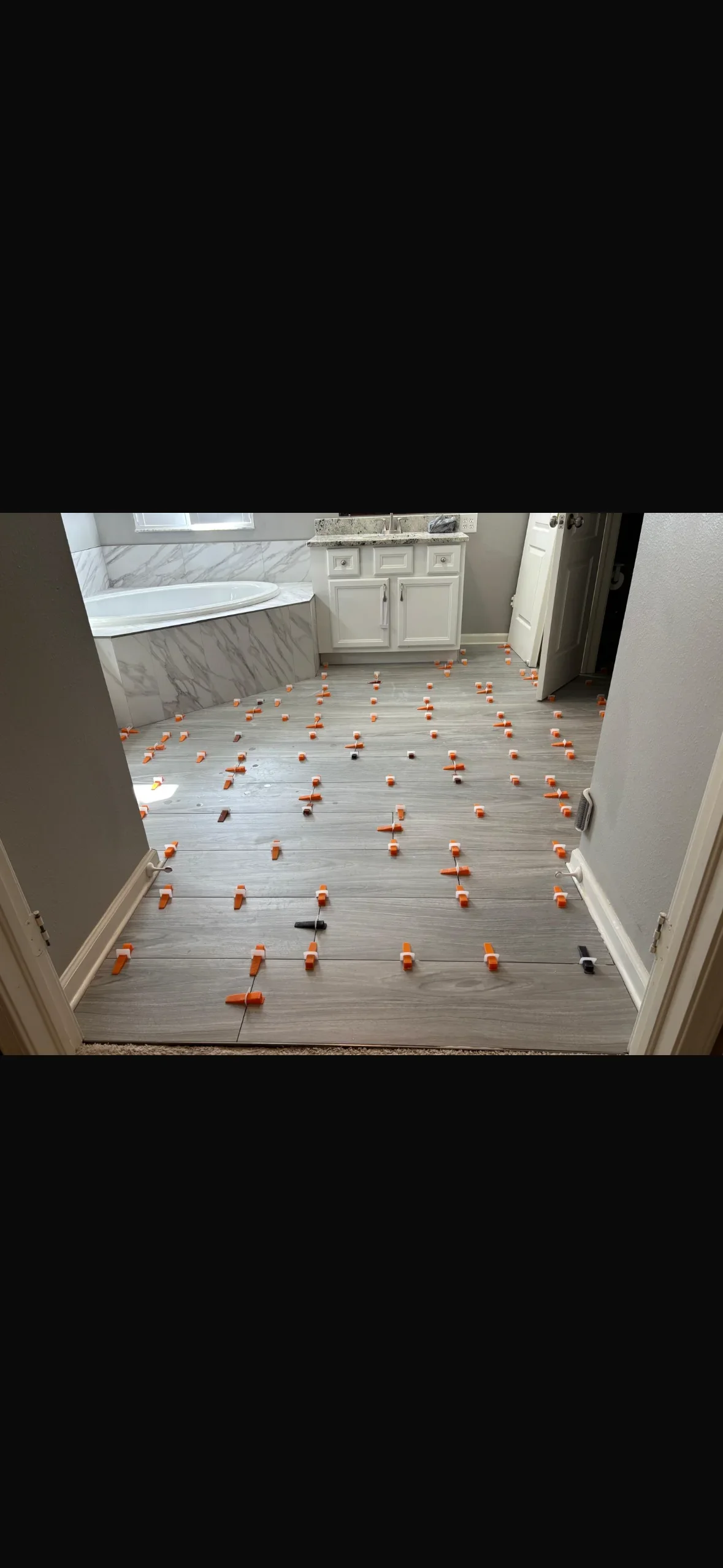 Gray wood-look porcelain floor tiles with orange leveling spacers in bathroom with white vanity and tub